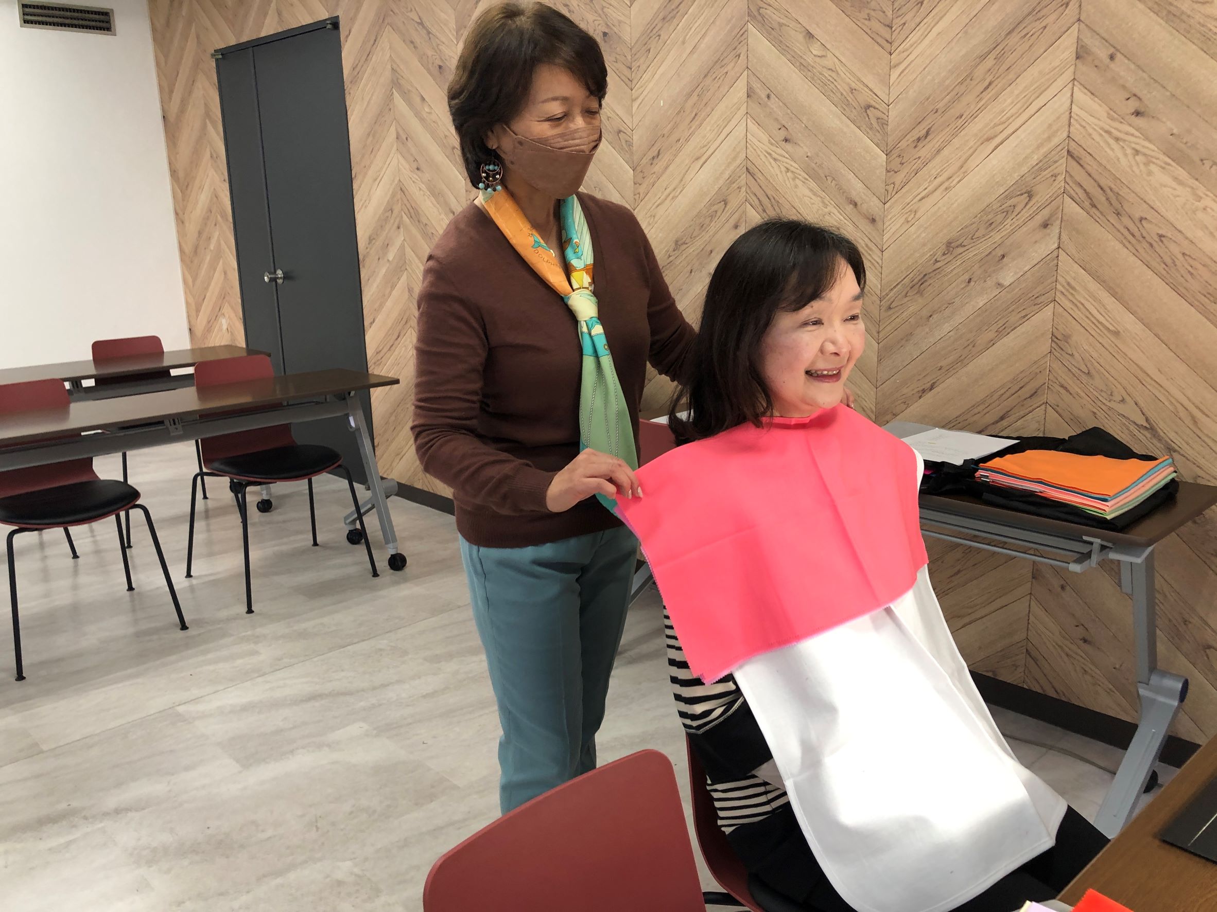 カラー診断を受ける参加社、先生が後ろに立ち参加社の顔の下に色の付いた布を拡げて鏡を見ながら説明しています。